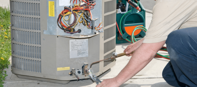 Technician performing maintenance on an outdoor air conditioning unit with tools and wiring visible