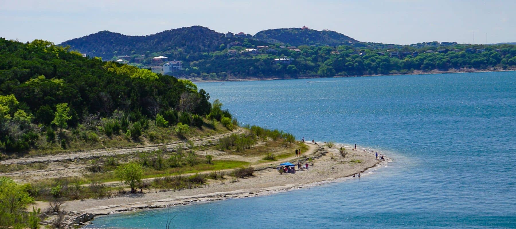 People enjoy a sunny day along the rocky shoreline of a lake surrounded by trees and distant hills