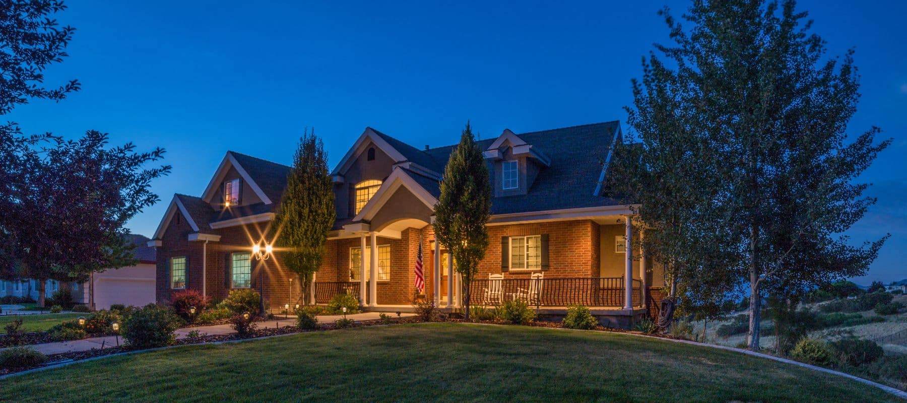 A warmly lit brick home with a front porch and landscaped yard at dusk under a clear blue sky