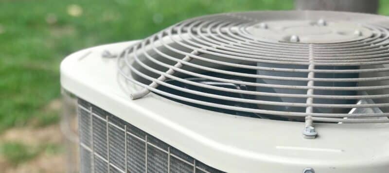 A close-up shot of the top of an outdoor air conditioning unit shows its fan grate and a glimpse of the fan blades, with a blurred green lawn in the background