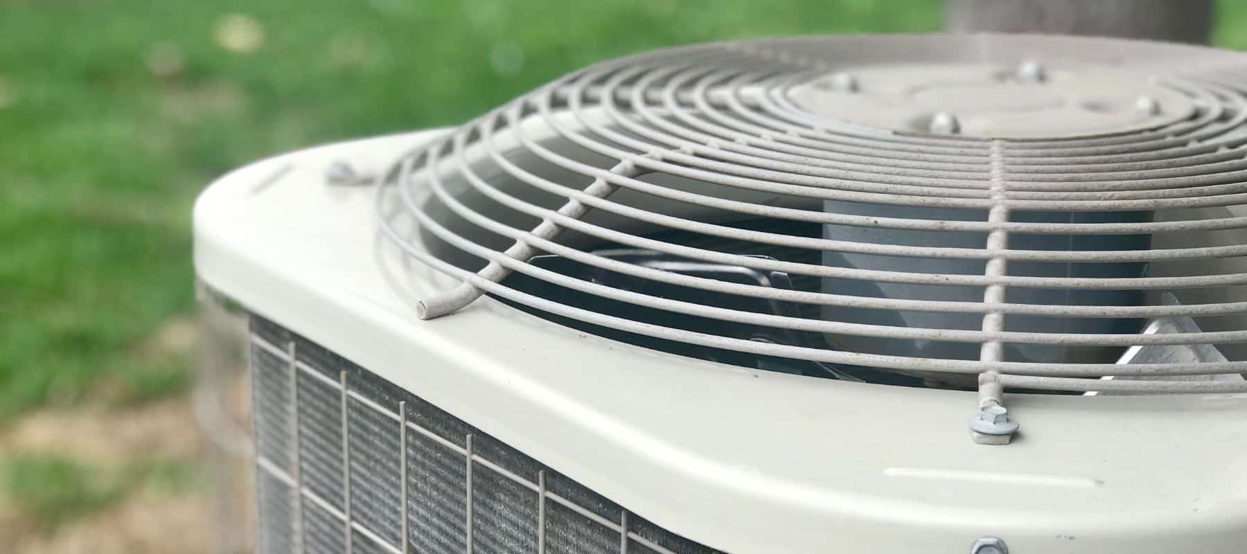 A close-up shot of the top of an outdoor air conditioning unit shows its fan grate and a glimpse of the fan blades, with a blurred green lawn in the background