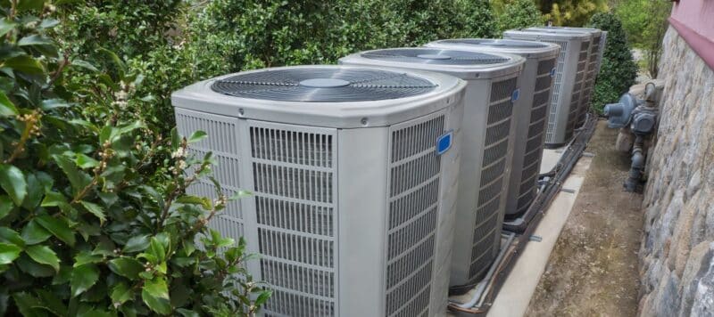 A row of multiple outdoor air conditioning units sits side-by-side on a concrete slab next to a green hedge and a stone wall