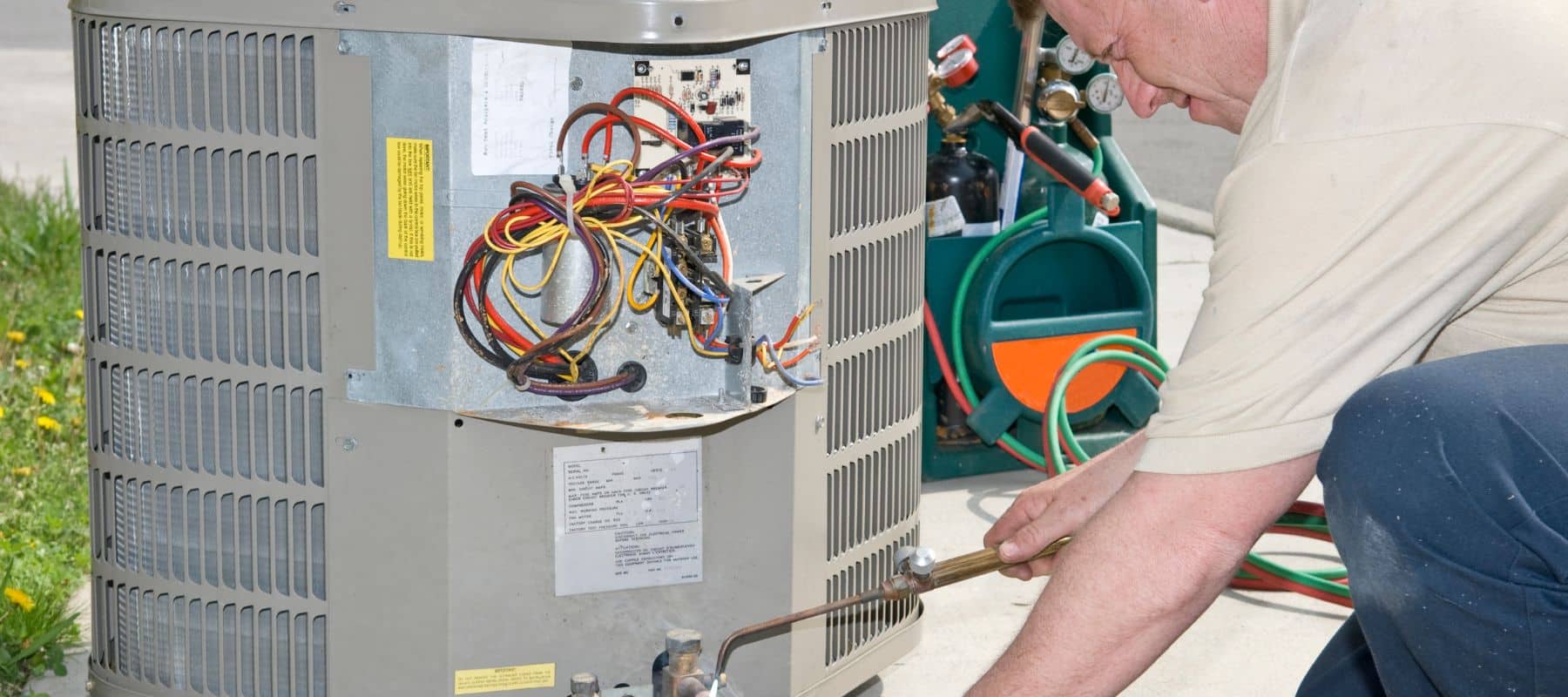 An HVAC technician works on an outdoor air conditioning unit with the panel open and wiring exposed